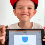 A boy smiling and holding an iPad showing his DoodleMaths Summer Challenge 2021 badge, which he's won by completing the Summer Challenge 2021